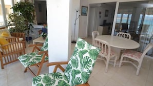 Uma área de refeições com uma mesa redonda e cadeiras, uma planta em vaso e vista para o mar através de uma porta de vidro.