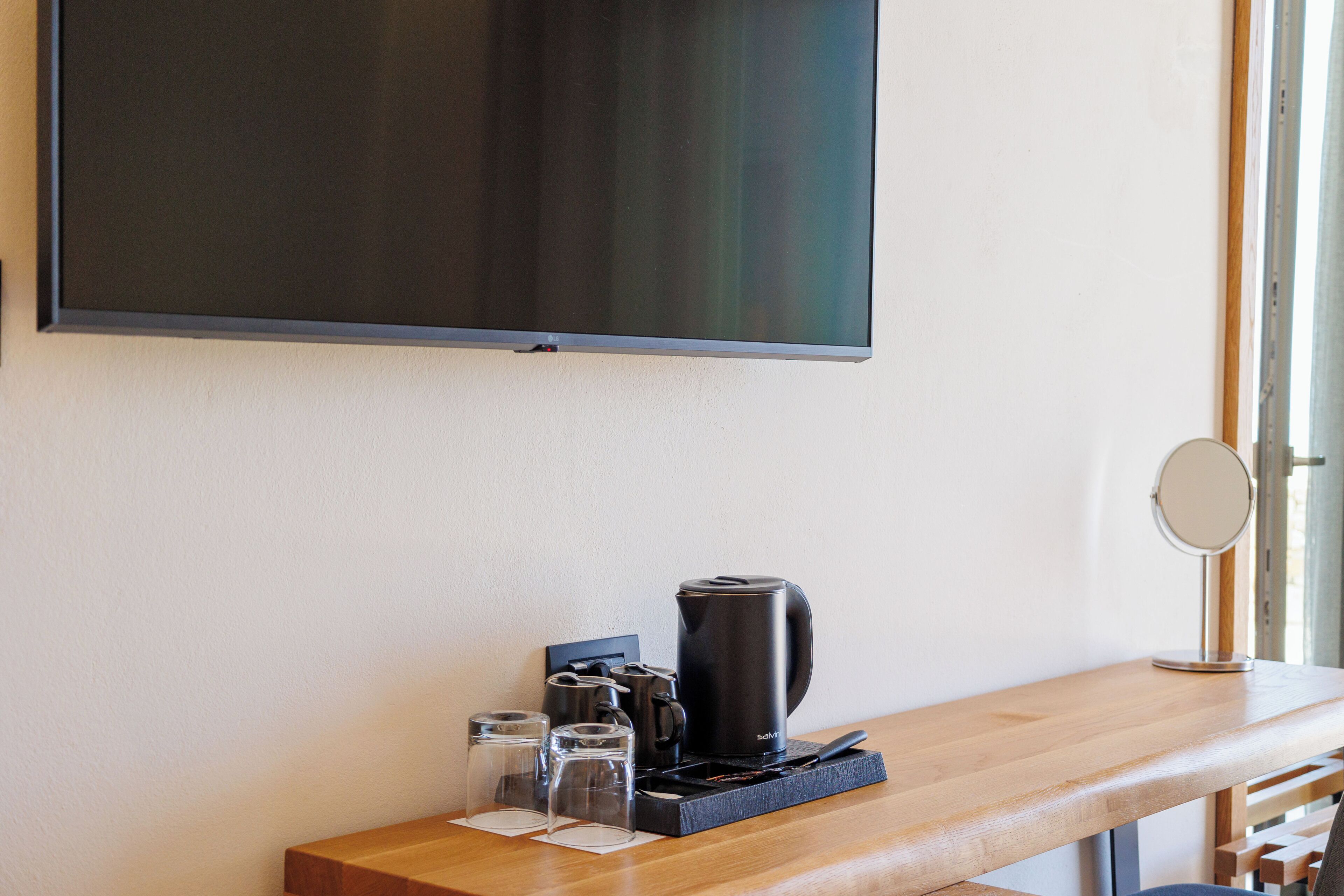 Une chambre d’hôtel dotée d’un téléviseur à écran plat fixé au mur, d’une table de chevet en bois, d’une cafetière, de verres et d’un petit miroir.
