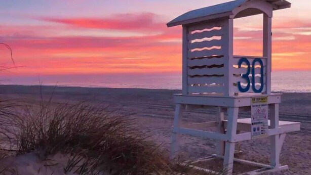 Une tour de surveillance, portant le numéro 30, située sur une plage bordée de dunes de sable, avec un coucher de soleil en arrière-plan.