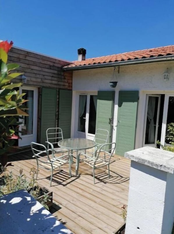 A patio with a round table and chairs, a wooden deck, and a building with green shutters and a tiled roof.