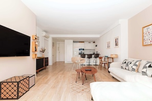 Uma sala de estar moderna com sofá, mesa de centro e televisão. Cozinha visível ao fundo, com área de jantar.