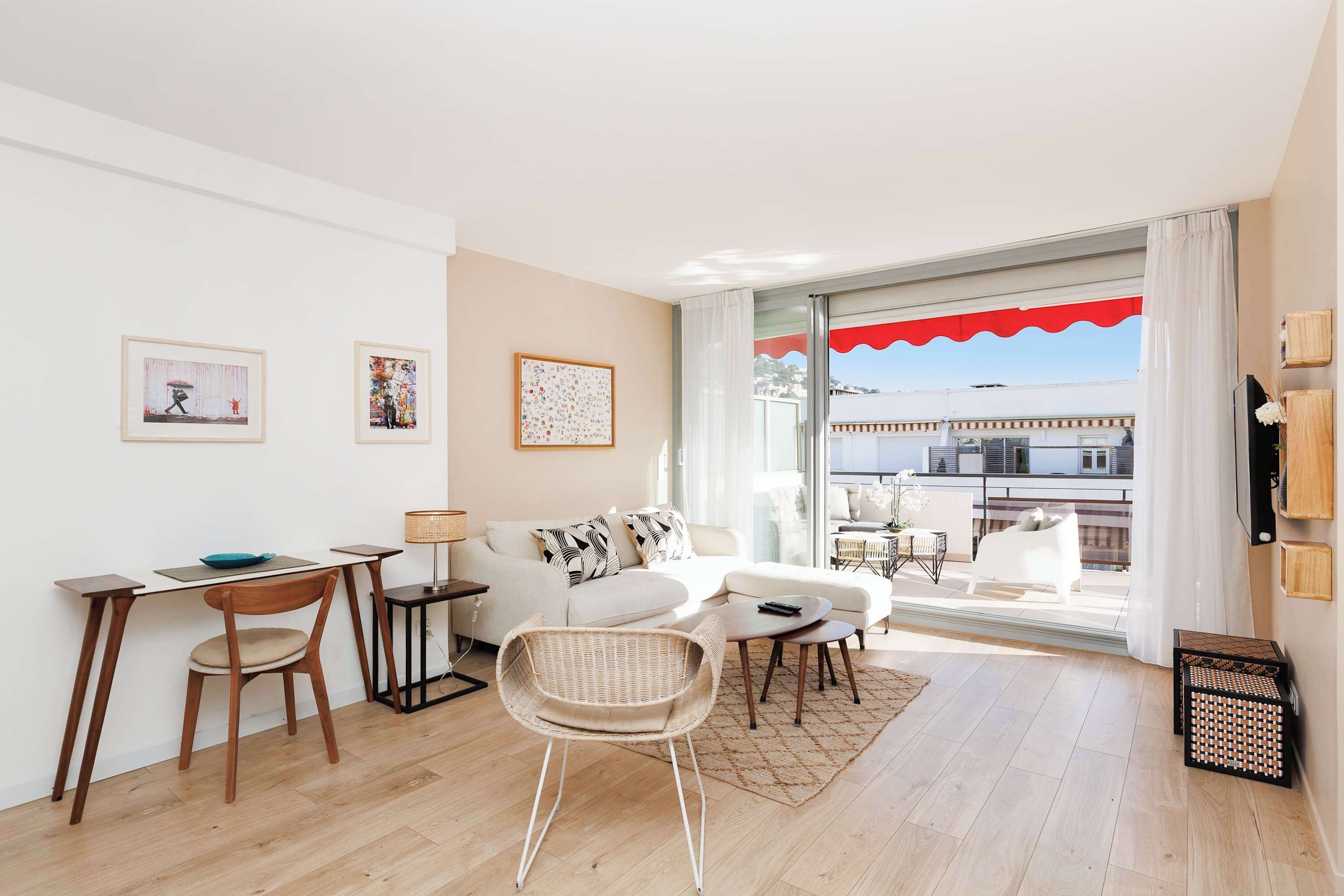 A modern living room with a dining area, a sofa, a wicker chair, a small table, and artwork on the wall.