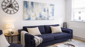 Un soggiorno con un divano blu scuro, un tavolino rotondo con una lampada, una sveglia a parete e un grande quadro astratto.