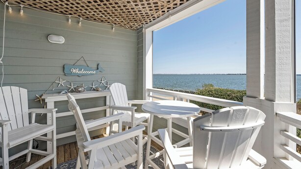 A covered outdoor seating area with white Adirondack chairs, a small round table, and a 'Whalecome' sign on the wall. - Captain's Quarters: Soundfront | Private Boat Slip (Manteo)