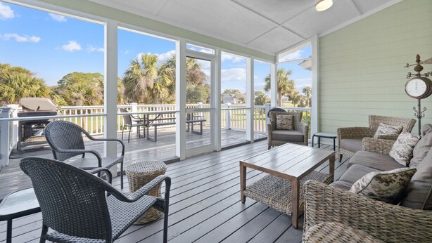 A covered outdoor deck with wicker furniture, a wooden table, and a view of palm trees and a house. - Sea Sounds -  steps to beach access, sleeps 17 and golf cart included (Fripp Island)