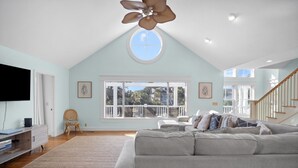 A spacious living room with a high ceiling, a large window, and a ceiling fan. - Sea Sounds -  steps to beach access, sleeps 17 and golf cart included (Fripp Island)