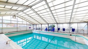 Piscina interna com paredes de vidro transparente, cadeiras azuis nas laterais e estrutura de telhado metálica.