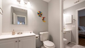 Un baño moderno con lavamanos, inodoro y tocador blancos. Se observan obras de arte decorativas en la pared y un espejo sobre el lavamanos.