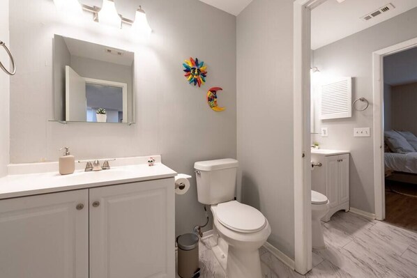 Un baño moderno con lavamanos, inodoro y tocador blancos. Se observan obras de arte decorativas en la pared y un espejo sobre el lavamanos.
