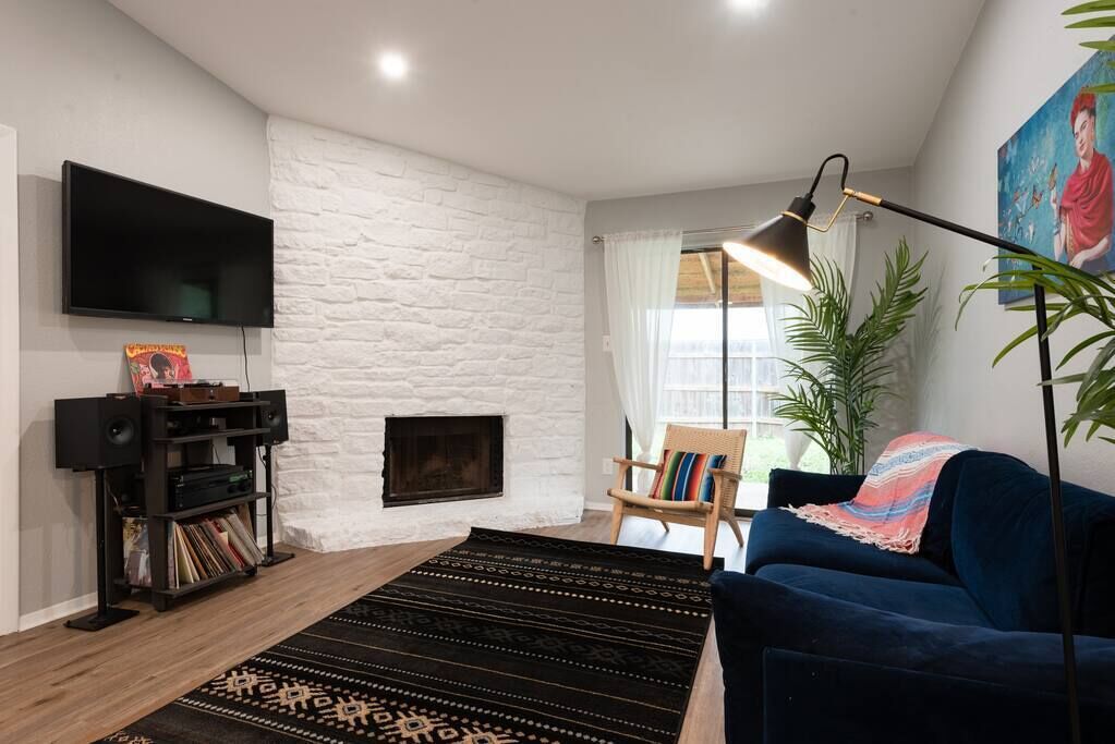 A living room with a white brick wall, a flat-screen TV, a fireplace, a blue sofa, a floor lamp, and a bookshelf.