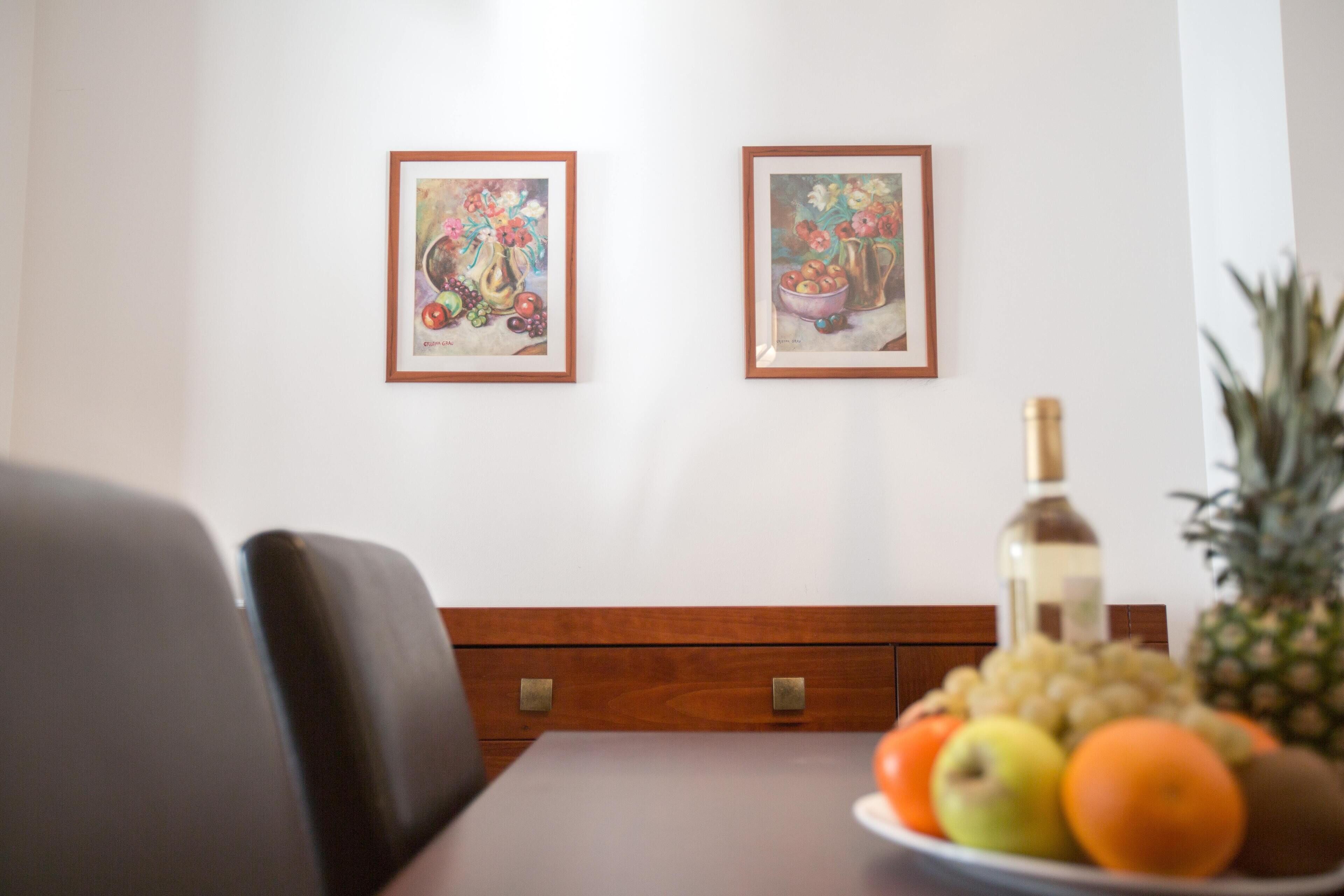 Ein Essbereich mit einem für zwei Personen gedeckten Tisch, mit Obstschale und Flasche. Über dem Tisch hängen zwei gerahmte Kunstwerke.