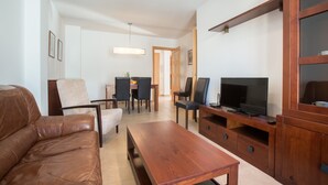 En moderne stue med en lædersofa, et træbord og et fjernsyn på et træstel. I baggrunden er der et spiseområde med stole og et bord.