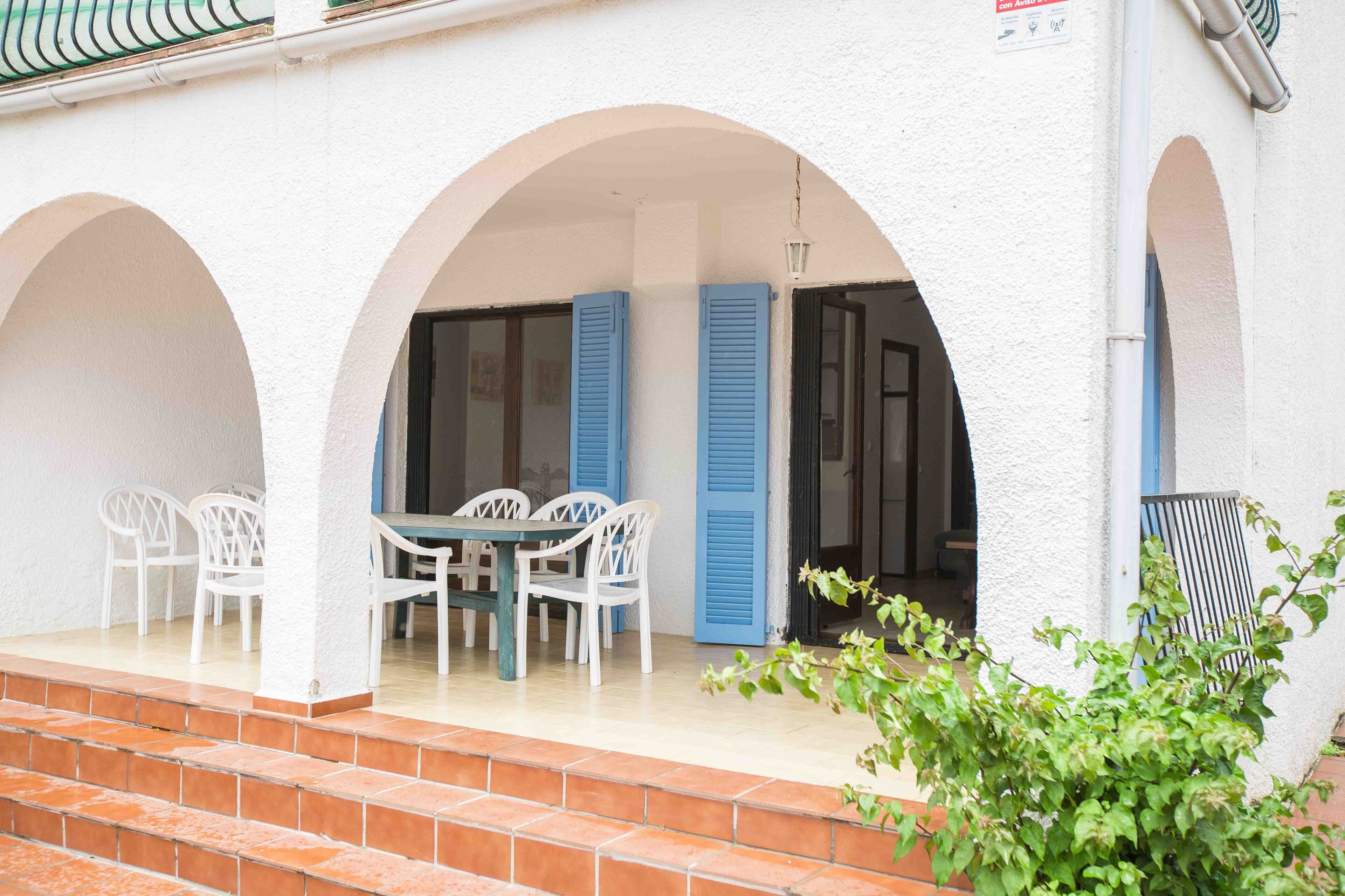 Une terrasse couverte avec une table et des chaises, des murs blancs et des volets bleus.