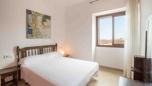 A bedroom with a bed, a nightstand, a painting, a window, and a view of the city.