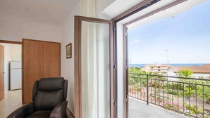A room with a sliding glass door leading to a balcony with a view of the sea and buildings.