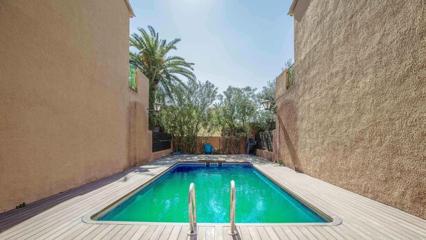 Ein rechteckiger Pool mit zwei Metallleitern auf einer Holzterrasse, umgeben von beigen Wänden und Palmen.