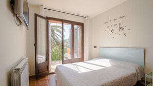 Ein Schlafzimmer mit Bett, Heizkörper, Fernseher und einem Balkon mit Blick auf Palmen.