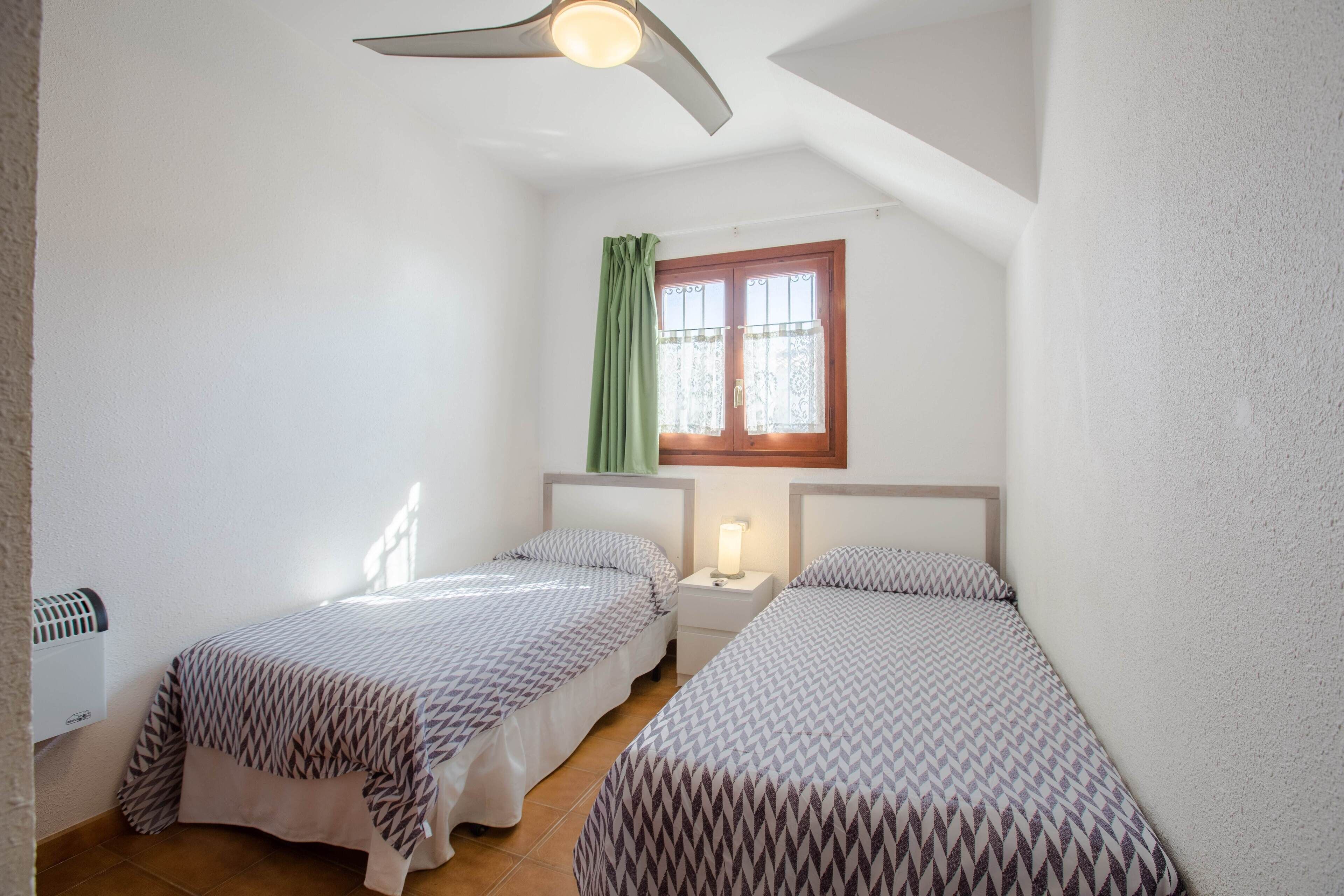 Un dormitorio pequeño y bien iluminado, con dos camas, una ventana con cortina y un ventilador de techo.
