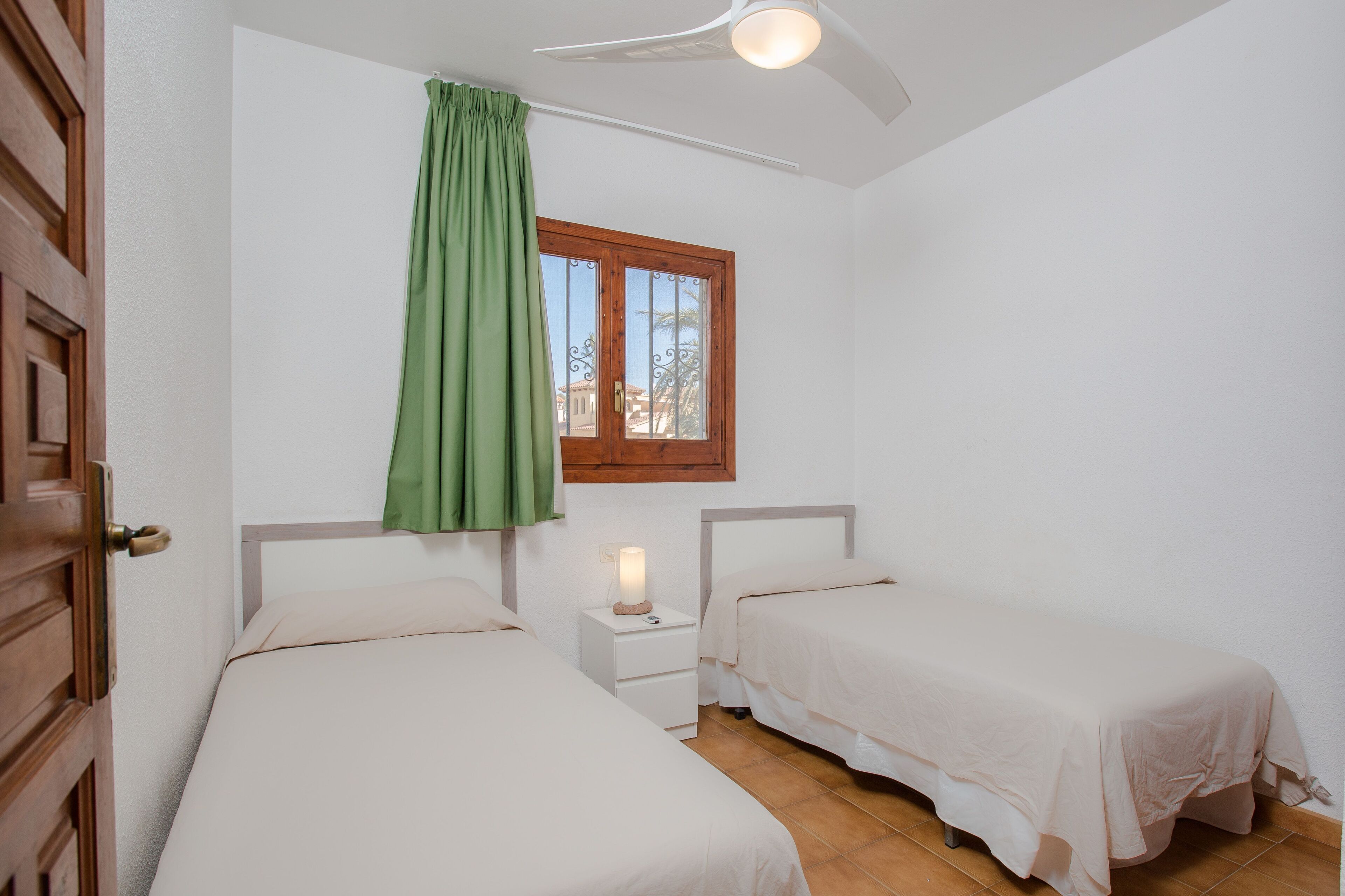 Una habitación con dos camas, una ventana con cortina, una mesita de noche con lámpara y un ventilador de techo.