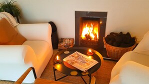 Un salon chaleureux avec une cheminée, des canapés blancs, une table basse avec un journal et un panier de bûches.