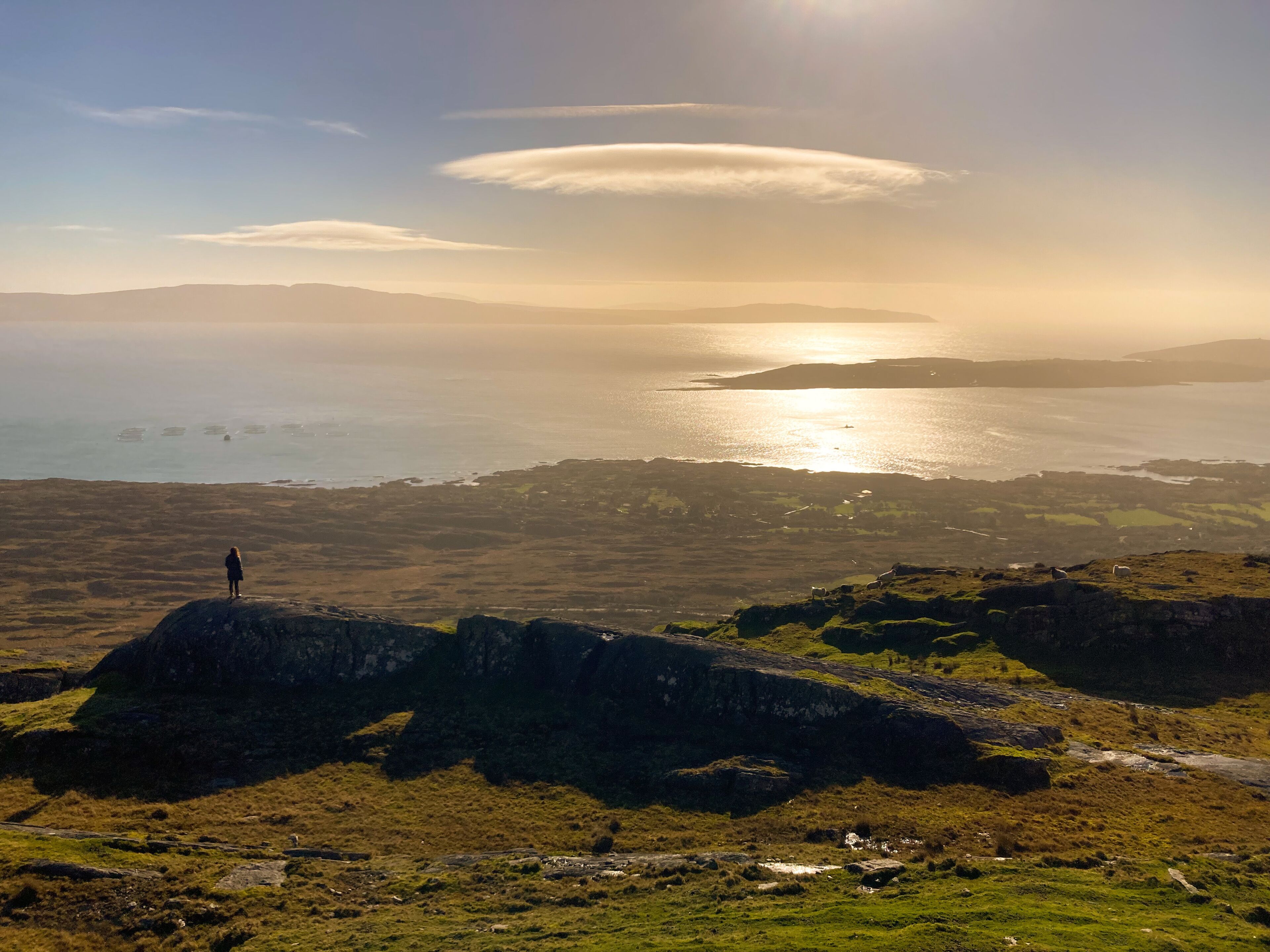 A person standing on a rocky outcrop overlooking a coastal landscape with a view of the sea and distant hills.
