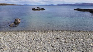 Une plage de galets bordée d’une eau claire, avec des montagnes à l’horizon et un ciel partiellement nuageux.