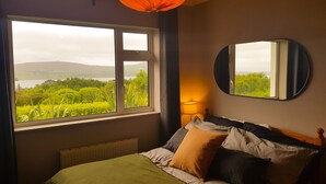 Une chambre à coucher avec un lit, une fenêtre donnant sur un paysage verdoyant et des collines, un miroir rond et une lampe.