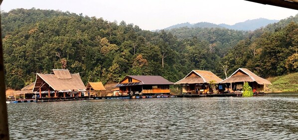 Floating houses with thatched roofs on a calm lake, surrounded by dense forest and hills. - Houseboat Floating, Enjoy the sounds of nature whit a unique place. (Tambon Ban Pao)