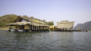 A floating house with a thatched roof and a wooden deck, situated on calm water with a forested hillside in the background.