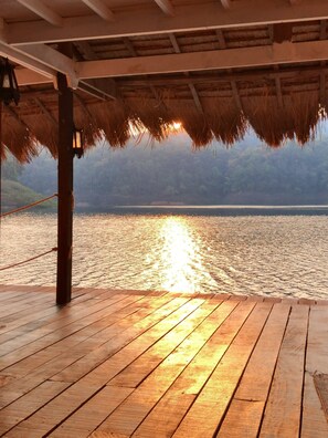 A wooden deck with a thatched roof, overlooking a calm body of water with sunlight reflecting on the surface. - Houseboat Floating, Enjoy the sounds of nature whit a unique place. (Tambon Ban Pao)