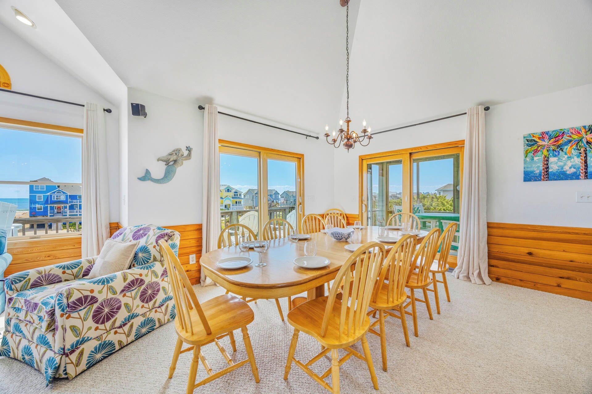 A dining area with a wooden table and chairs, a sofa with a floral pattern, and a view of the beach through large windows.