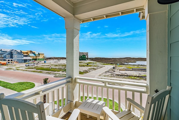 A porch with white railings and a table, overlooking a beach area with buildings and a road.