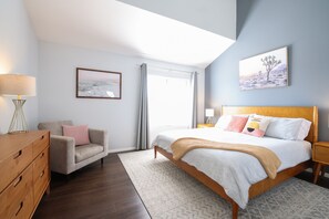 Um quarto com cama, mesa de cabeceira, penteadeira, cadeira e janela com cortinas.