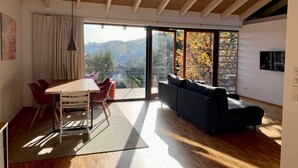 Amplia sala de estar con techo de madera, una mesa de comedor con sillas, un sofá y puertas de vidrio correderas que dan a un balcón con vistas a árboles y colinas.