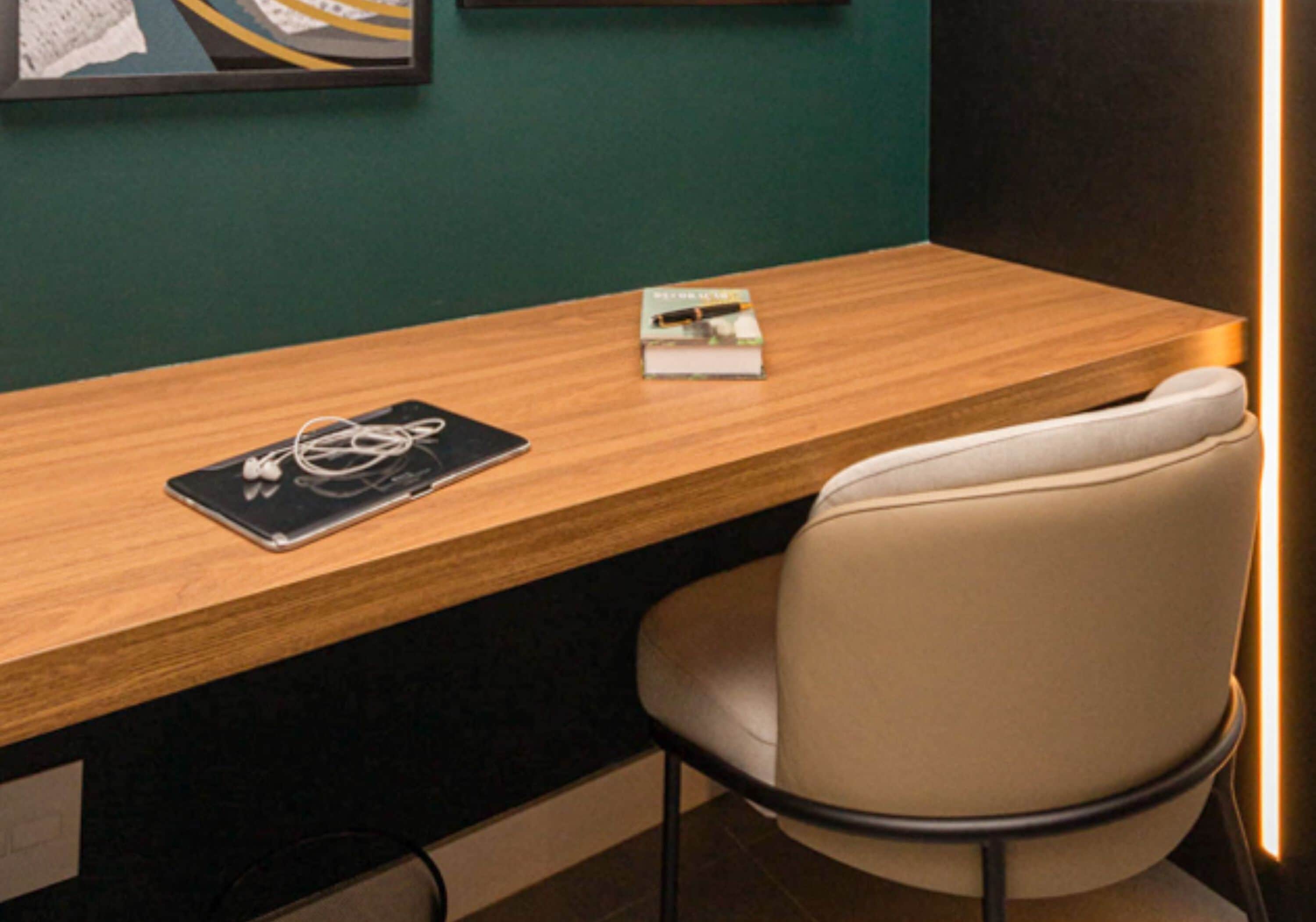 A wooden desk with a laptop and a book, a chair, and a wall with a framed picture.