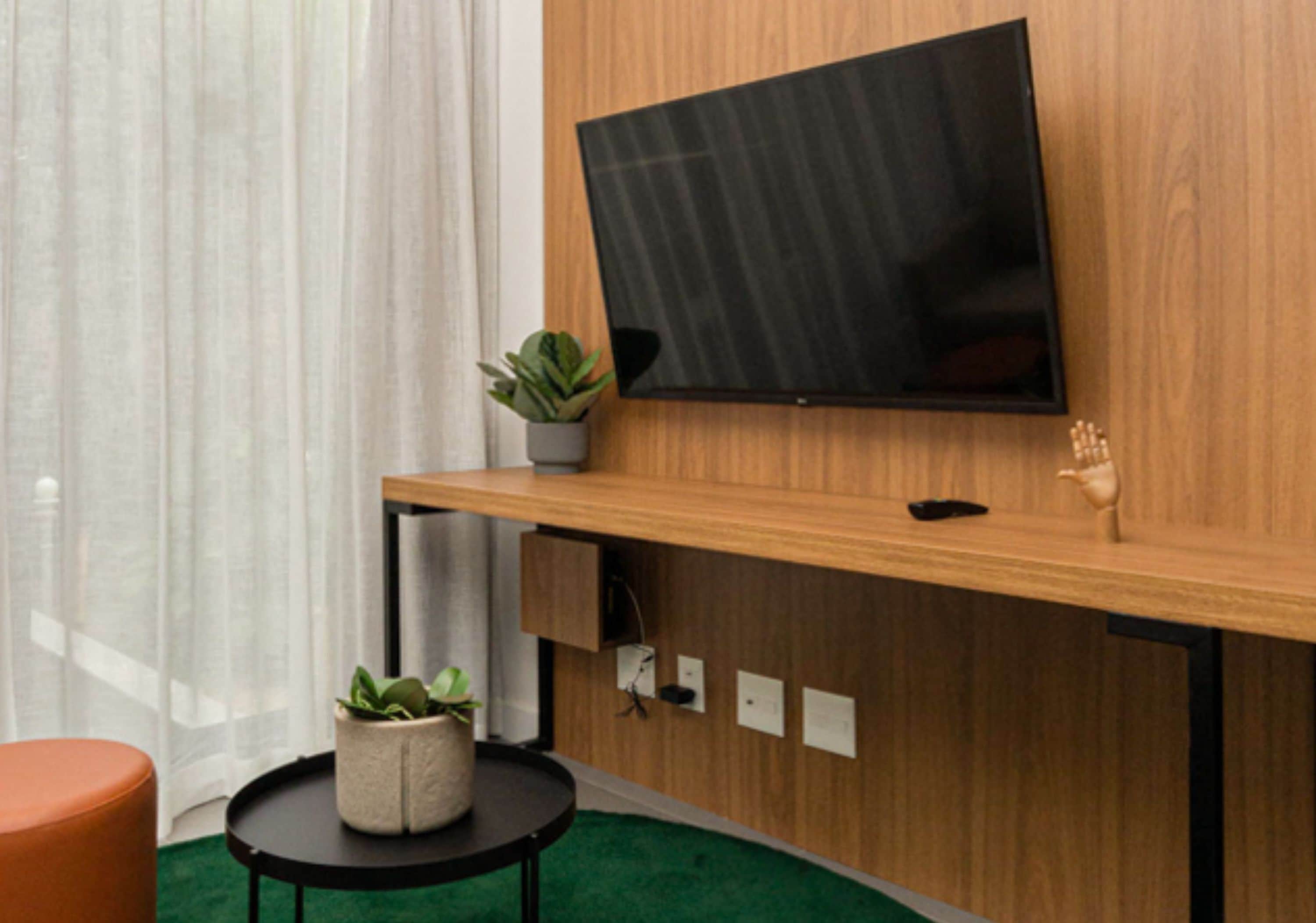 A modern room with a wooden wall-mounted TV, a small potted plant, a round table with a cushion, and a green carpet.