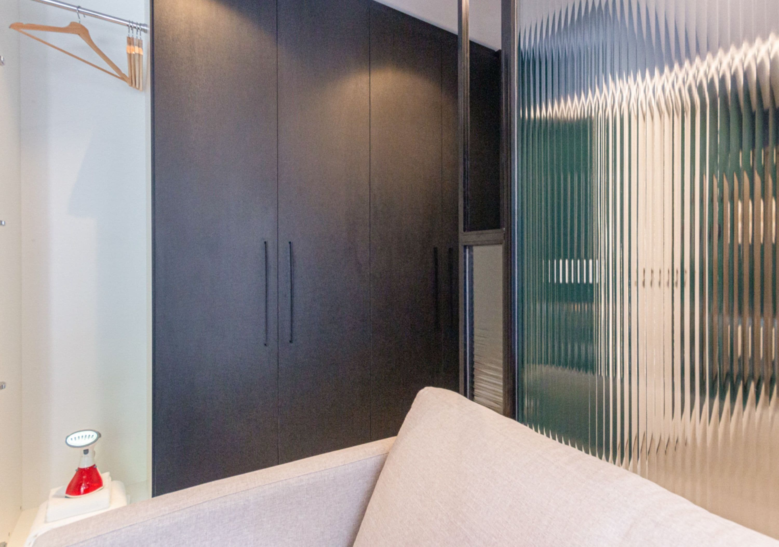 A modern bedroom with a dark wardrobe, a white bed, and a glass partition with vertical blinds.