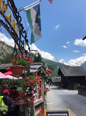 A rustic wooden building with a mountain backdrop, a flag, and a flower-adorned window.