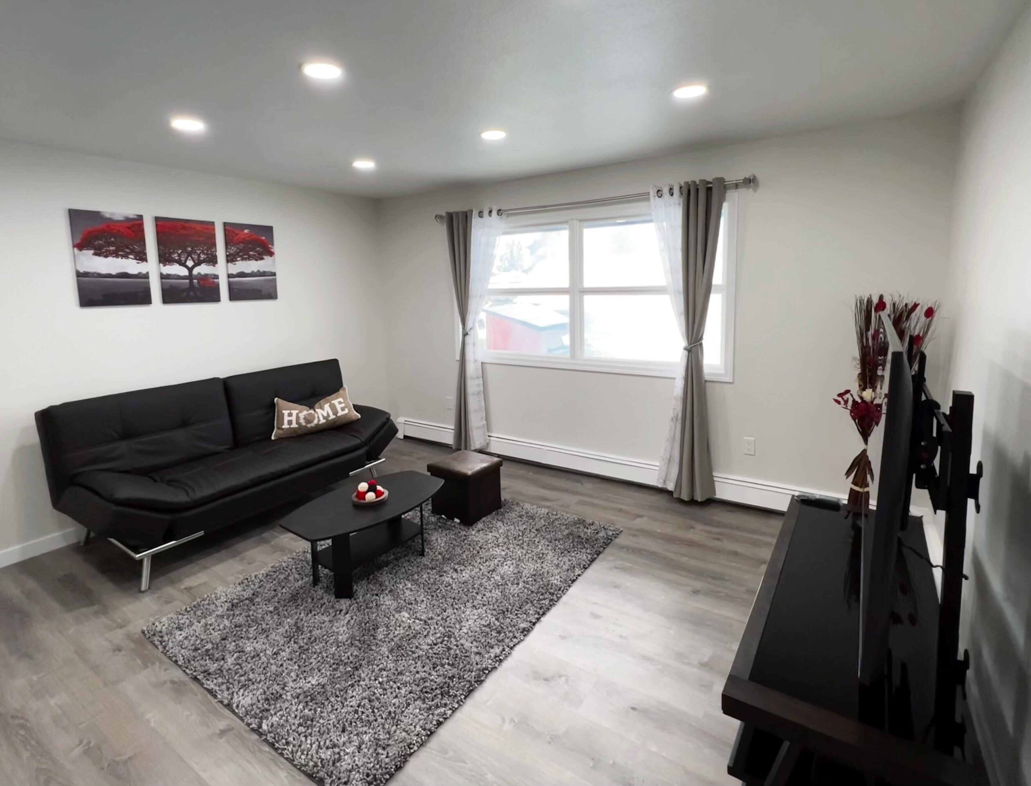 A modern living room with a black sofa, a coffee table, and a TV stand. There are three wall-mounted picture frames and a window with curtains.
