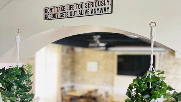 Un comedor interior con follaje colgante y un cartel que dice "DON'T TAKE LIFE TOO SERIOUSLY. NOBODY GETS OUT ALIVE ANYWAY".