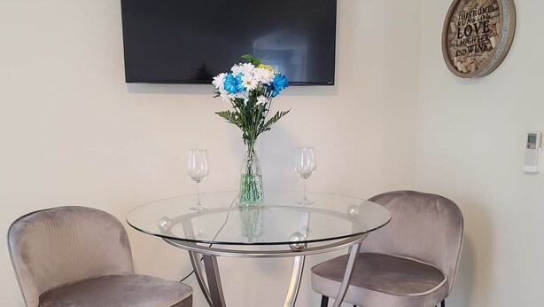 Un coin repas avec une table en verre, deux chaises, un vase de fleurs, un téléviseur et une assiette décorative accrochée au mur.