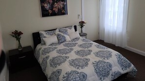 Une chambre à coucher avec un lit, une table de chevet, un tableau accroché au mur et une fenêtre avec des rideaux.