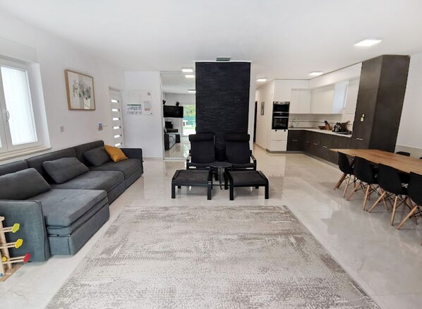 Un salon moderne avec un canapé gris, des chaises noires et une table basse. On y trouve également un coin cuisine avec des meubles blancs et une table à manger avec des chaises.