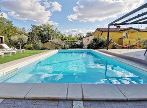 Une piscine rectangulaire dont les bords sont recouverts de carreaux, entourée d’une terrasse dallée. On y trouve des chaises longues et un hamac.