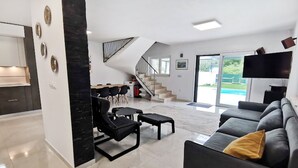 Un salon moderne avec un canapé noir, une table basse et un coin repas avec des chaises. Un escalier mène à un balcon offrant une vue sur la piscine.