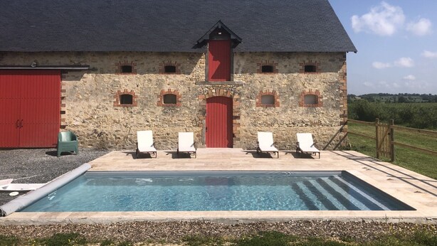 A stone building with a red door and windows, a swimming pool, and white lounge chairs.