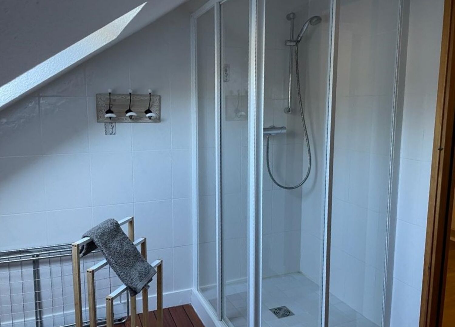 A shower with a glass door, a towel rack with three towels, and a wooden chair with a towel.