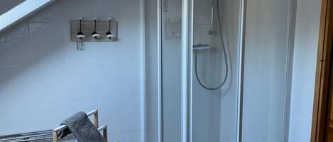 A shower with a glass door, a towel rack with three towels, and a wooden chair with a towel.