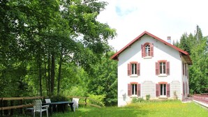 Une maison blanche aux volets rouges, entourée d’une clôture en bois et d’un espace verdoyant avec une table et des chaises.