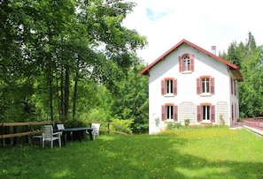 A white house with red shutters, surrounded by a wooden fence and a grassy area with a table and chairs.
